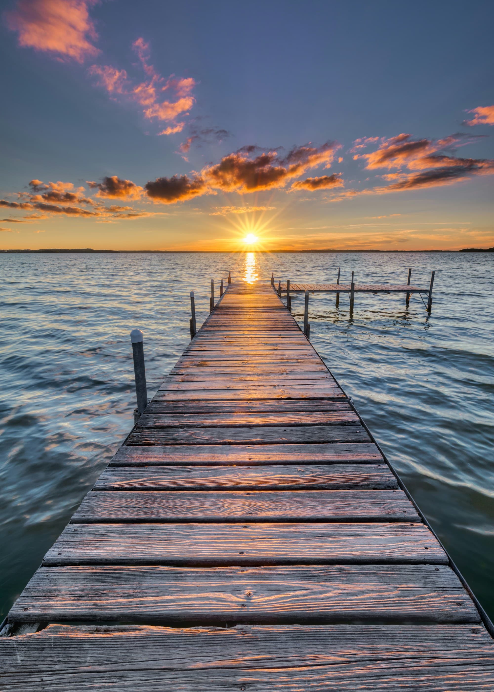 A wooden dock reaching out to a golden sunburst sunset over a calm lake
